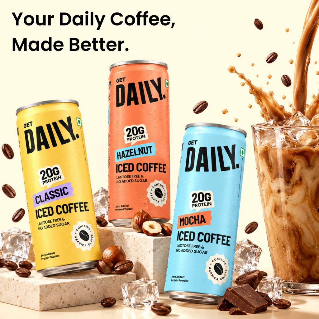Three Get Daily. 20g protein iced coffee cans in yellow, orange, and blue, displayed with coffee beans, chocolate chunks, ice cubes, and a glass of iced coffee with a coffee splash against a light beige background.